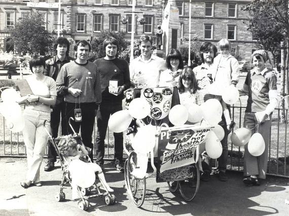 A black and white photo of a group of people standing in a city square holding balloons. There is a child's pram covered in protest signs and buttons.